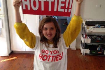 A young girl in a yellow onesie and a "Go You Hotties!" t-shirt smiling while holding up a red sign that says "My Mum's a HOTTIE!!" in a home setting.