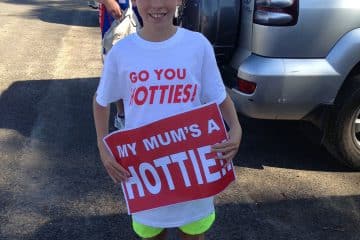 A young boy in a "Go You Hotties!" t-shirt smiling while holding a red "My Mum's a HOTTIE!!" sign in a parking lot