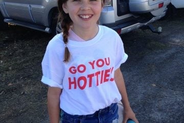 A young girl with a side braid smiling and wearing a white "Go You Hotties!" t-shirt and denim shorts