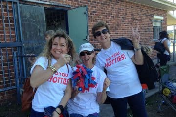 Three women wearing matching "Go You Hotties!" t-shirts smiling and cheering with red and blue pom-poms in front of a brick building.