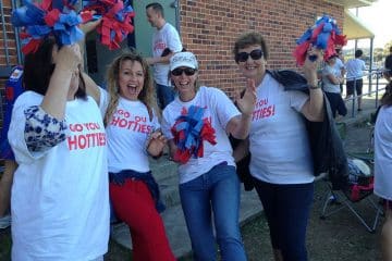 Four women wearing matching "Go You Hotties!" t-shirts smiling and waving red and blue pom-poms while cheering in front of a brick building.