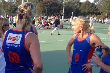 Two netball players in blue and red Hogan Hot Water sponsored uniforms standing on an outdoor court during a match break