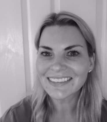 A black and white portrait of a smiling woman with light-coloured hair, looking directly at the camera.