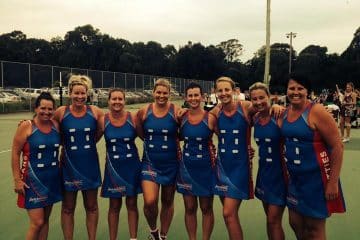 A group of eight women in blue and red netball uniforms smiling and posing together on an outdoor court under a cloudy sky.