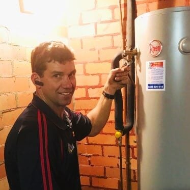 A technician in a dark blue polo shirt smiling while adjusting a valve on a tall, silver Rheem hot water heater