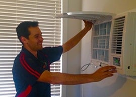 Home » Hot water system A technician in a dark blue polo with red stripes smiling while lifting the front cover to inspect the filter of a wall-mounted air conditioning unit.
