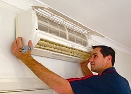 Home » Hot water system A technician in a dark blue polo with red stripes using both hands to carefully position a white, wall-mounted air conditioning unit against a ceiling.