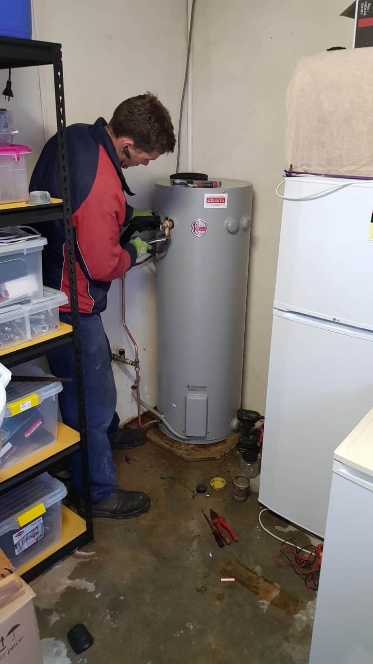 A technician in a red and black jacket using tools to work on the plumbing connections of a grey Rheem electric hot water storage tank installed in an indoor utility space.