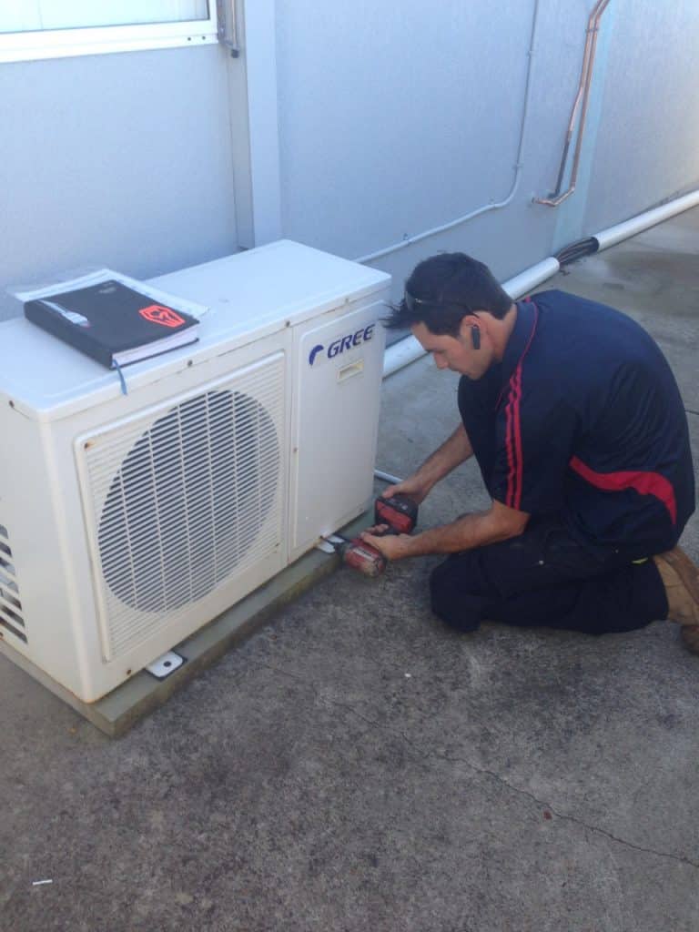A technician in a navy and red polo shirt kneeling on a concrete surface while using a power drill to secure a white Gree air conditioning condenser unit to its base.