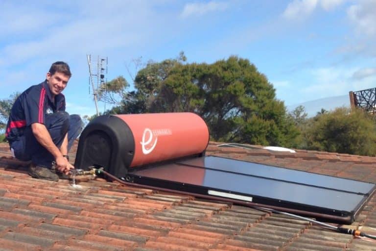 A smiling technician crouching on a tiled roof while performing maintenance on an Edwards solar hot water system