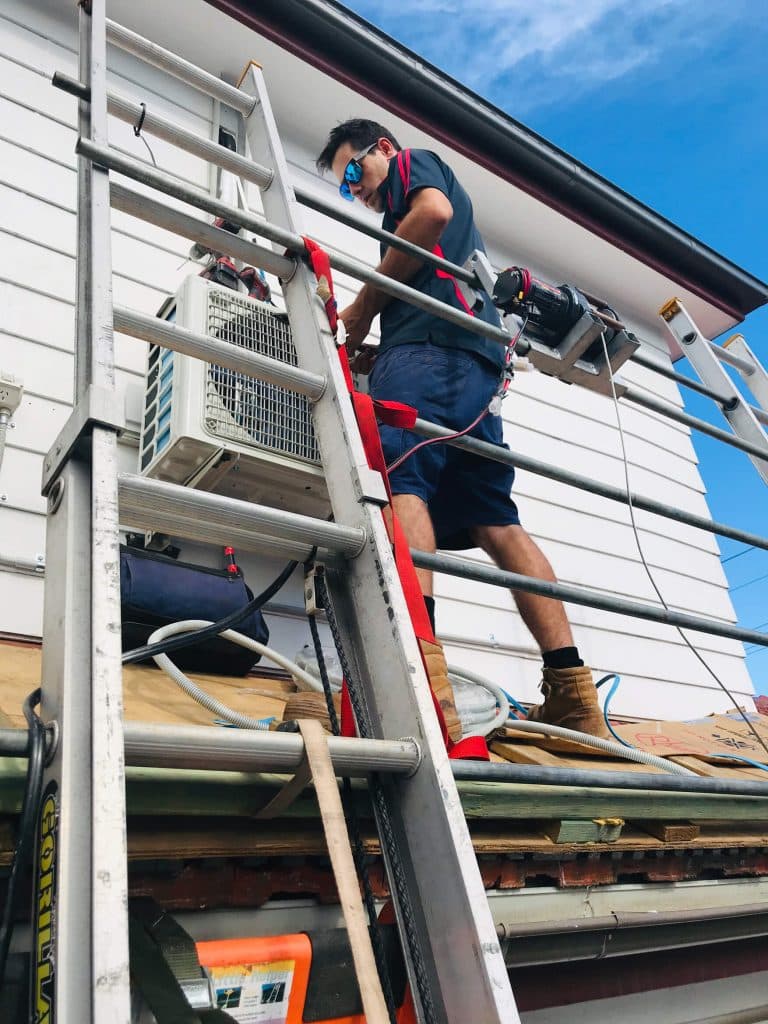 A low-angle shot of a technician standing on a roof while installing an air conditioning condenser unit on a white weatherboard wall