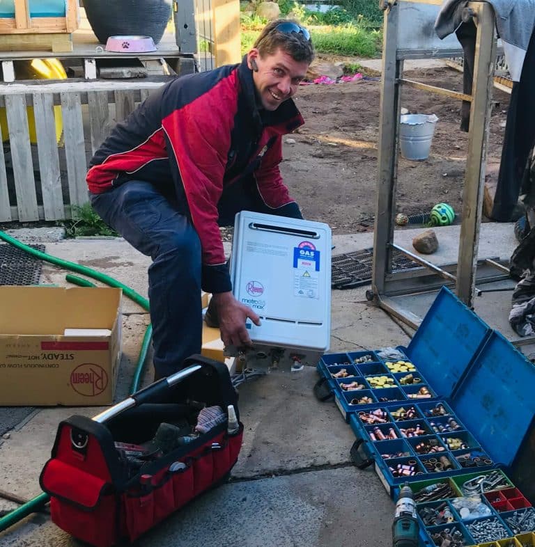 A smiling technician in a red and black jacket kneeling outdoors, preparing to install a white Rheem Metro continuous flow gas hot water unit