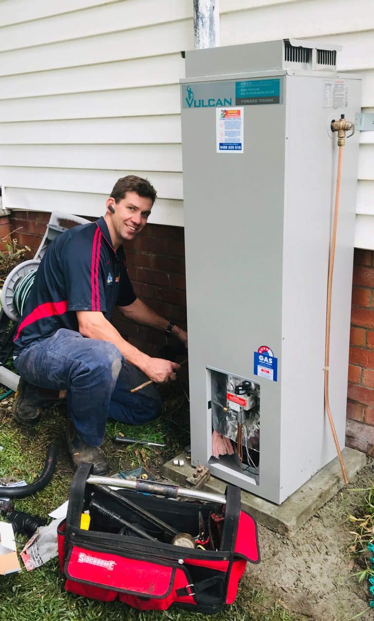 Mike kneeling next to a tall grey Vulcan "Freeloader" gas hot water storage tank during an outdoor installation