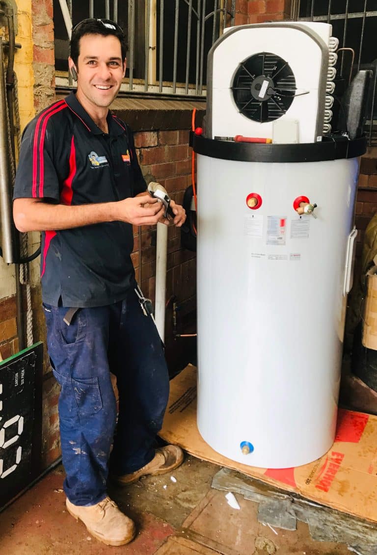 A smiling technician in a navy and red polo shirt stands next to a tall, white Bosch heat pump hot water system during installation