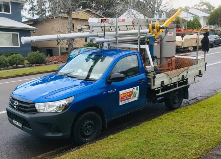 A blue Toyota Hilux service truck parked on a residential street, featuring a Hogan Hot Water & Air Conditioning logo on the door