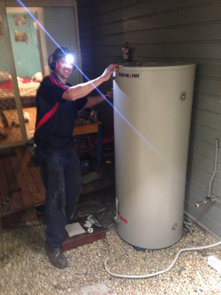 A smiling technician wearing a headlamp and earmuffs stands next to a tall, cream-coloured Thermann hot water storage tank
