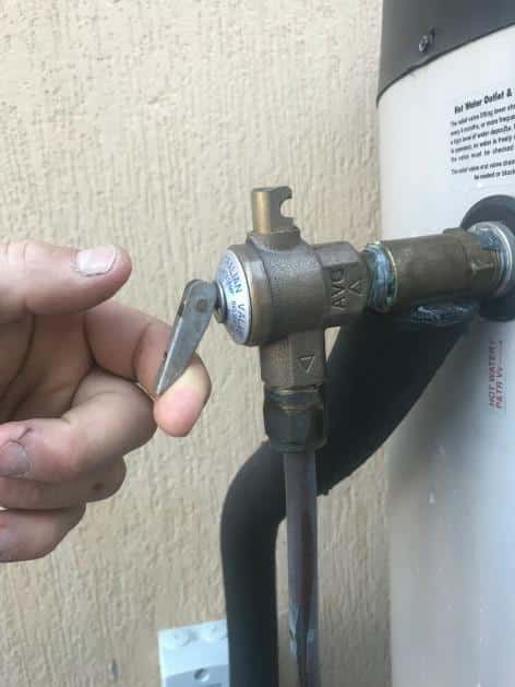A close-up of a hand gently lifting the small silver lever on a brass temperature and pressure relief (TPR) valve to test it