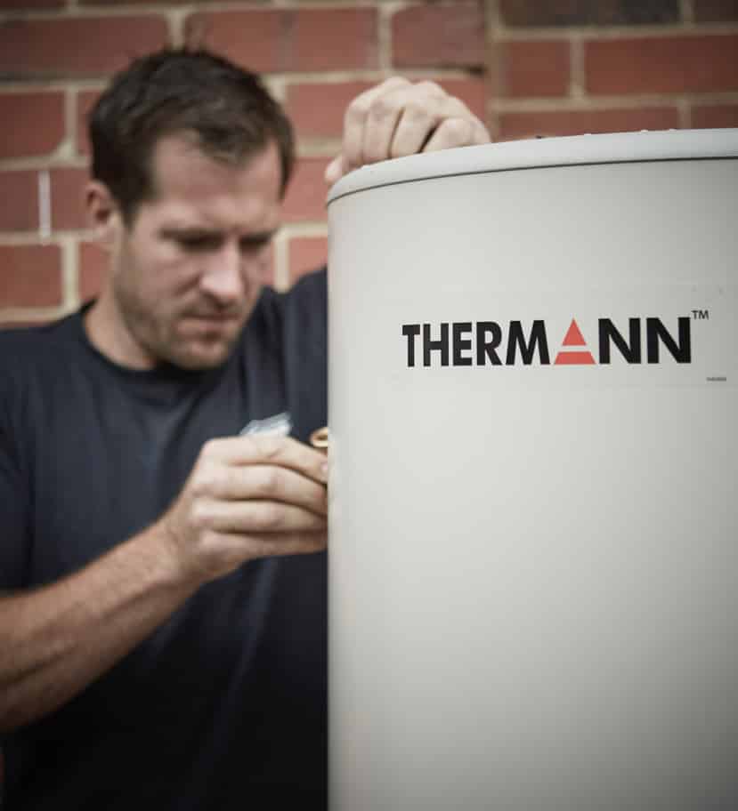 A technician in a dark shirt working on the side of a white Thermann brand water heater against a brick wall.