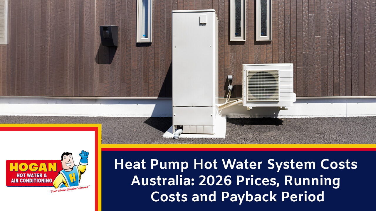 An outdoor heat pump hot water system with a tall white storage tank and an adjacent compressor unit installed against a brown slatted wall.