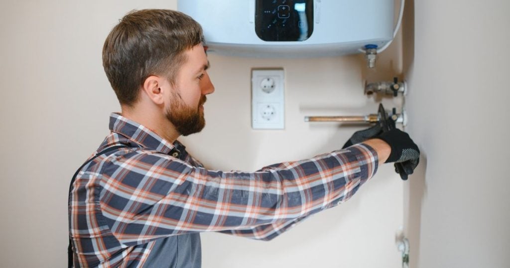 A technician uses a tool to work on the wall-mounted plumbing underneath an electric water heater.