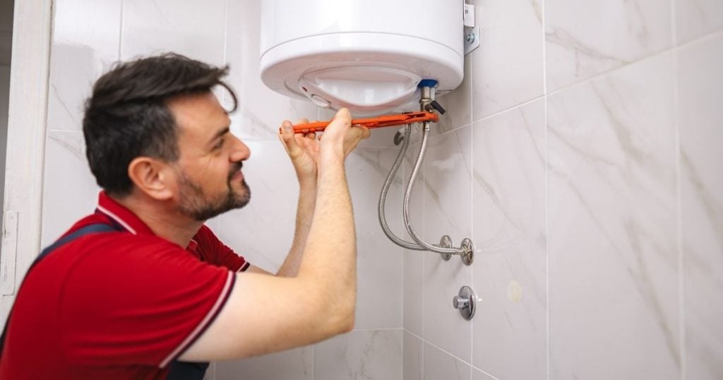 A technician uses an orange wrench to tighten the plumbing connections of a wall-mounted electric water heater.