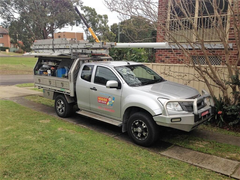 Hot Water Installation and Maintenance Lakelands » Lakelands A silver Isuzu utility vehicle parked on a driveway, featuring a "Hogan Hot Water" logo on the door and a specialised service body loaded with ladders, tools, and a plumbing pipe.
