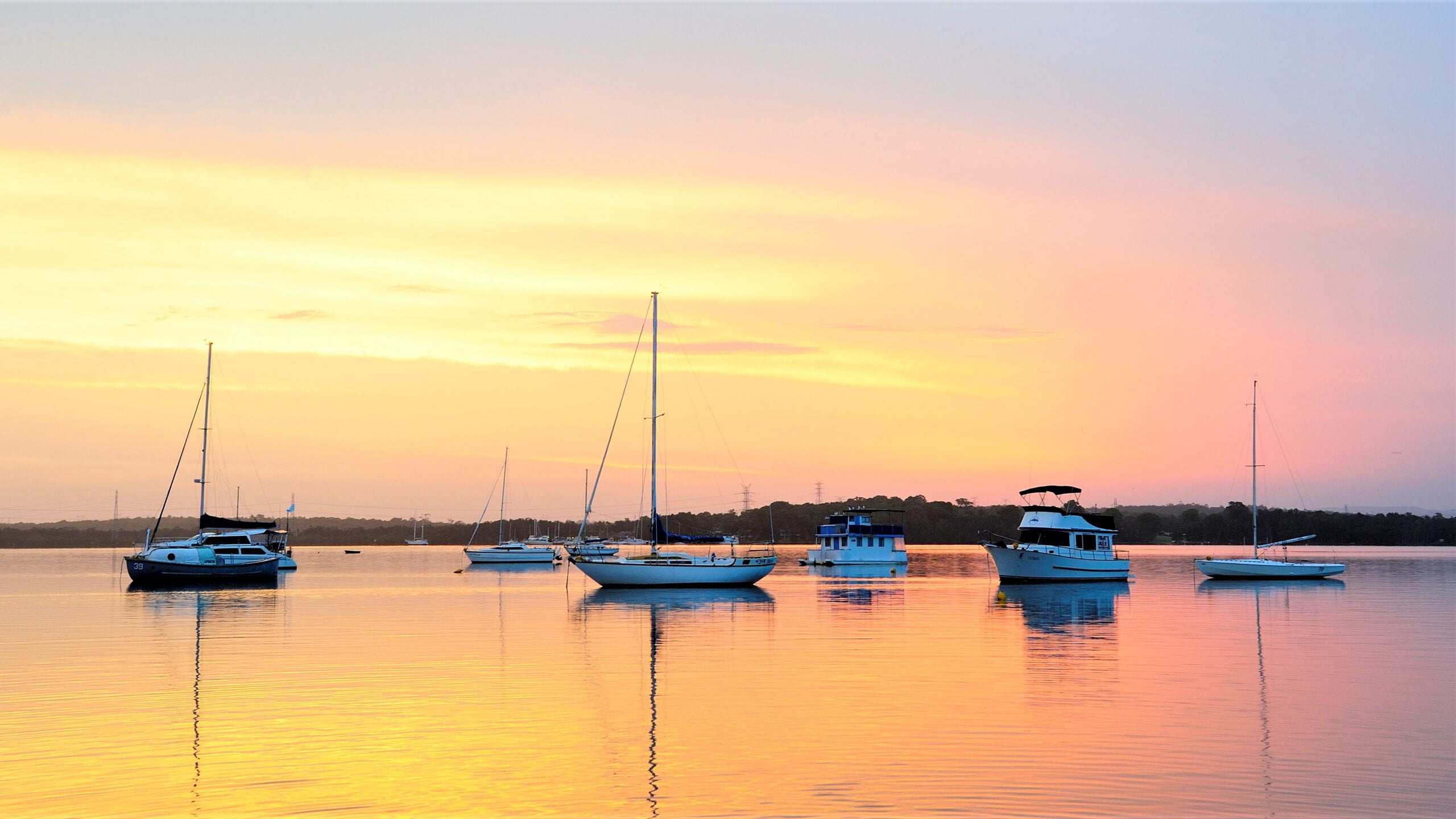 Several sailboats and motorboats anchored on a glassy, still lake during a pastel sunset.