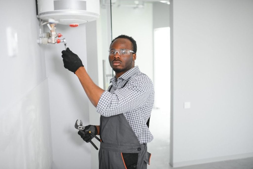 A technician wearing safety glasses, a plaid shirt, and grey overalls uses tools to adjust the water valves.