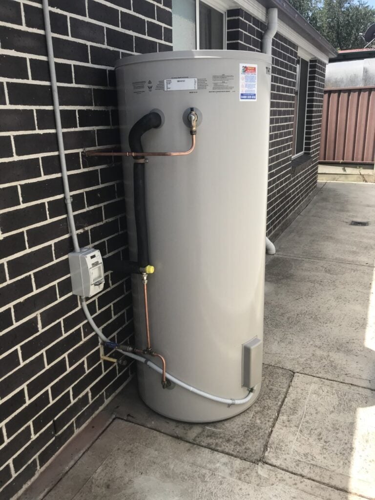 Outdoor hot water heater tank installed against a dark brick wall on a concrete pathway.