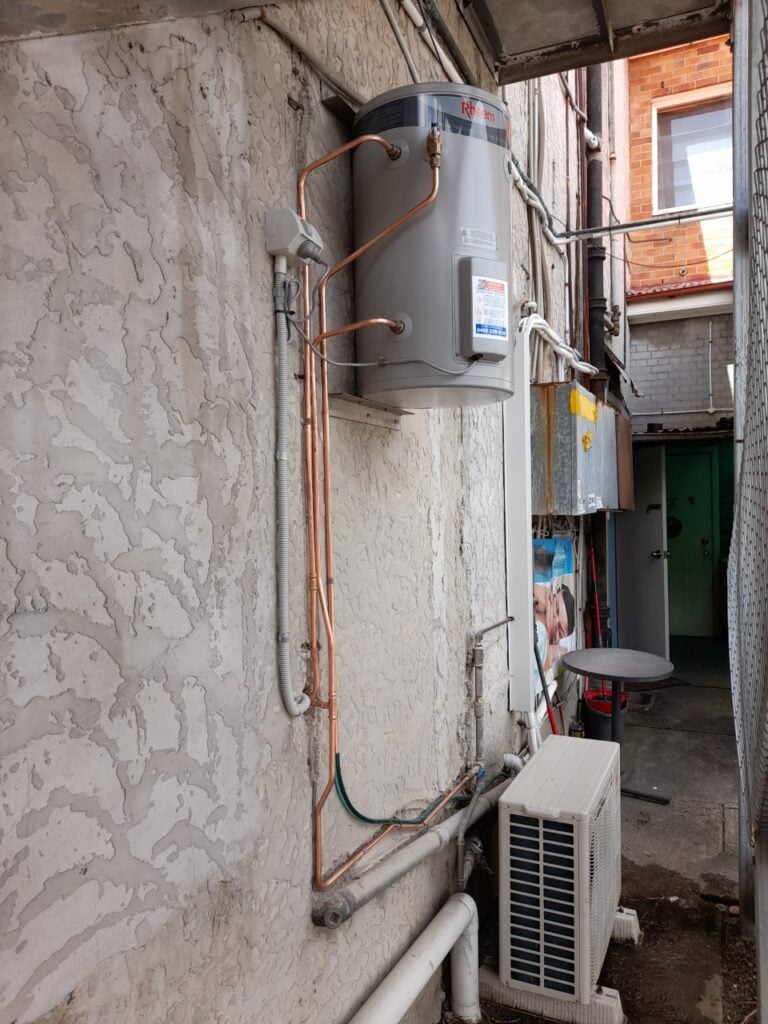 Wall-mounted Rheem electric water heater on a textured exterior wall with exposed copper piping.