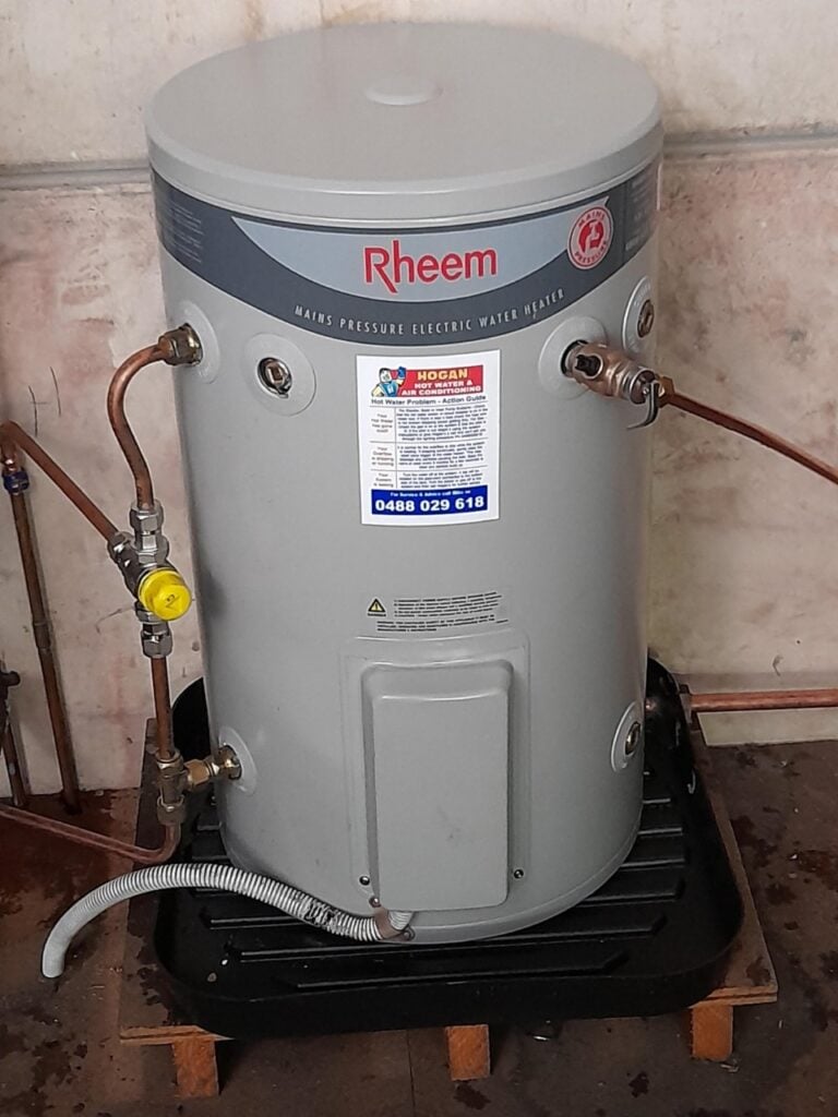 A grey Rheem electric water heater resting on a black drain pan with copper pipe fittings.