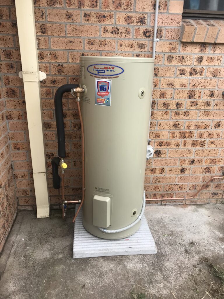 An AquaMax electric water heater set on a concrete pad against a brick house wall.