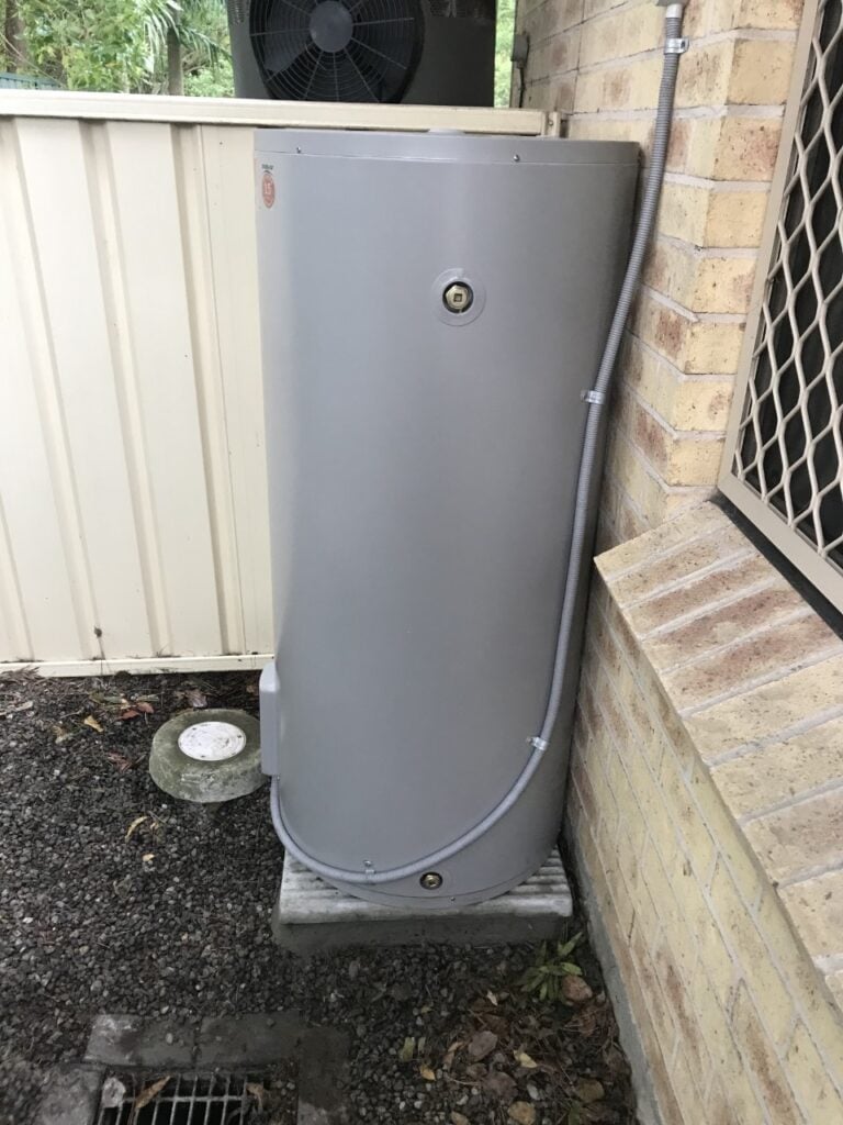Grey outdoor electric hot water tank installed on a base beside a brick wall.