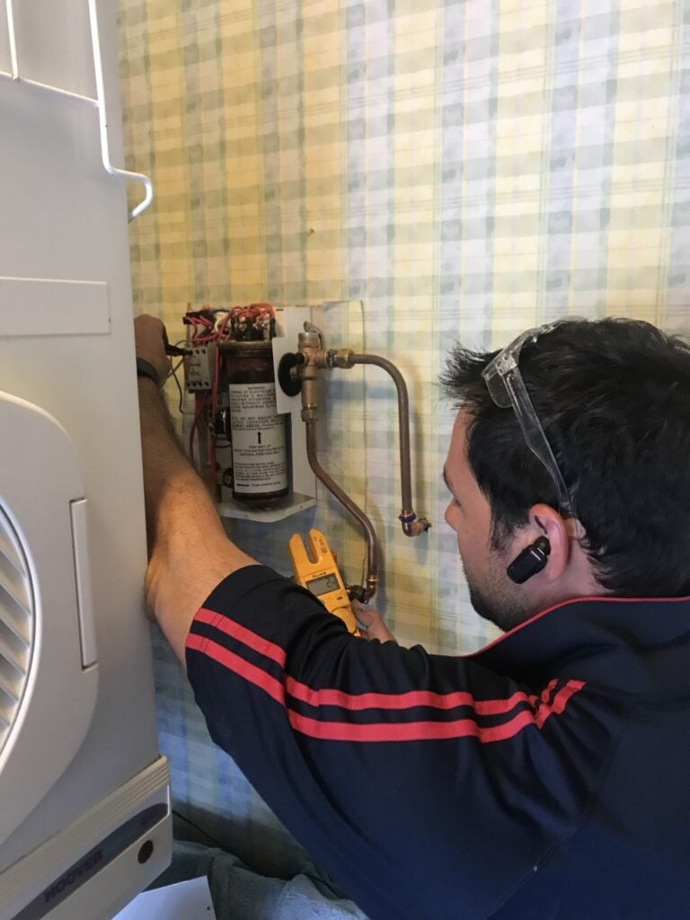 A technician wearing safety glasses uses a Fluke digital multimetre to test the electrical components of an indoor water heating unit mounted on a patterned wall.
