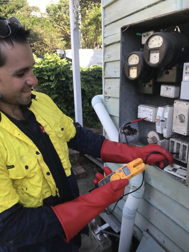 Technician in high-visibility shirt and safety gloves using a multimetre to test an outdoor electrical metre box.