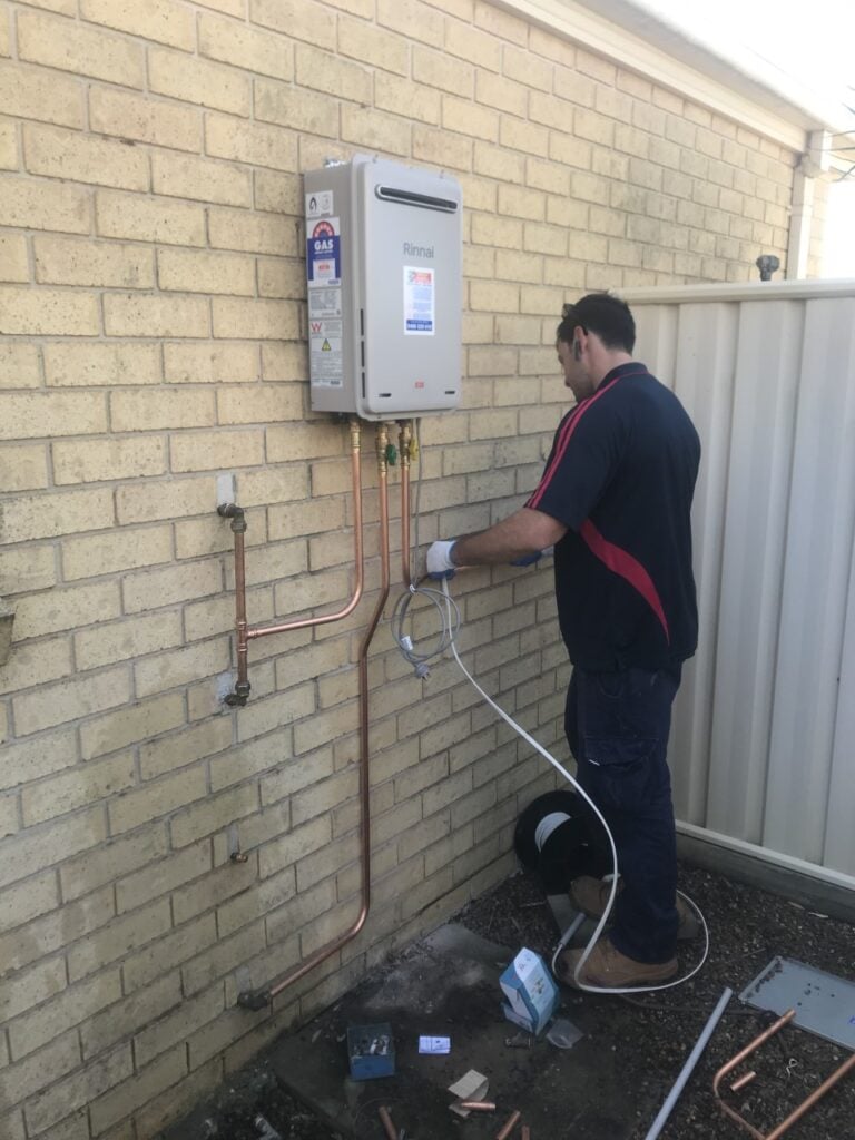 Technician installing wiring for a wall-mounted Rinnai continuous flow gas hot water system against a brick wall.