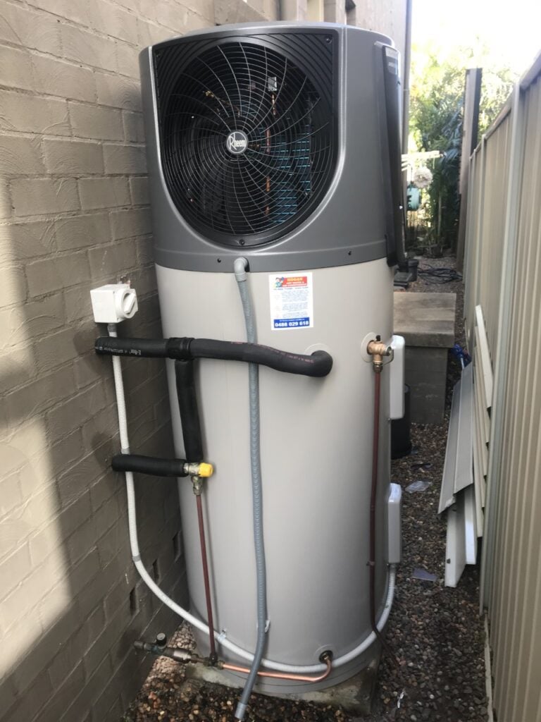 Outdoor Rheem heat pump hot water system installed in a narrow side path between a brick wall and a metal fence.