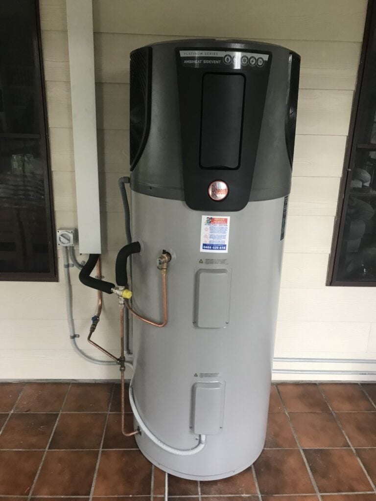 Rheem Ambiheat heat pump water heater installed on a tiled patio against a house wall.