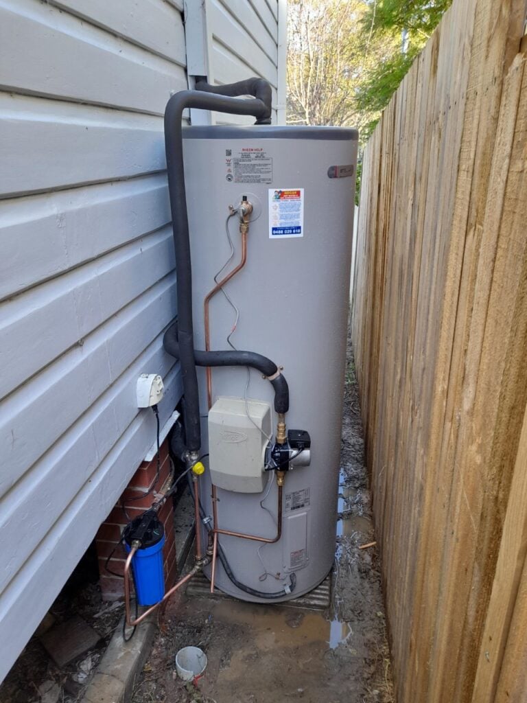 Outdoor grey water heater tank installed in a narrow alley between a house wall and a wooden fence.