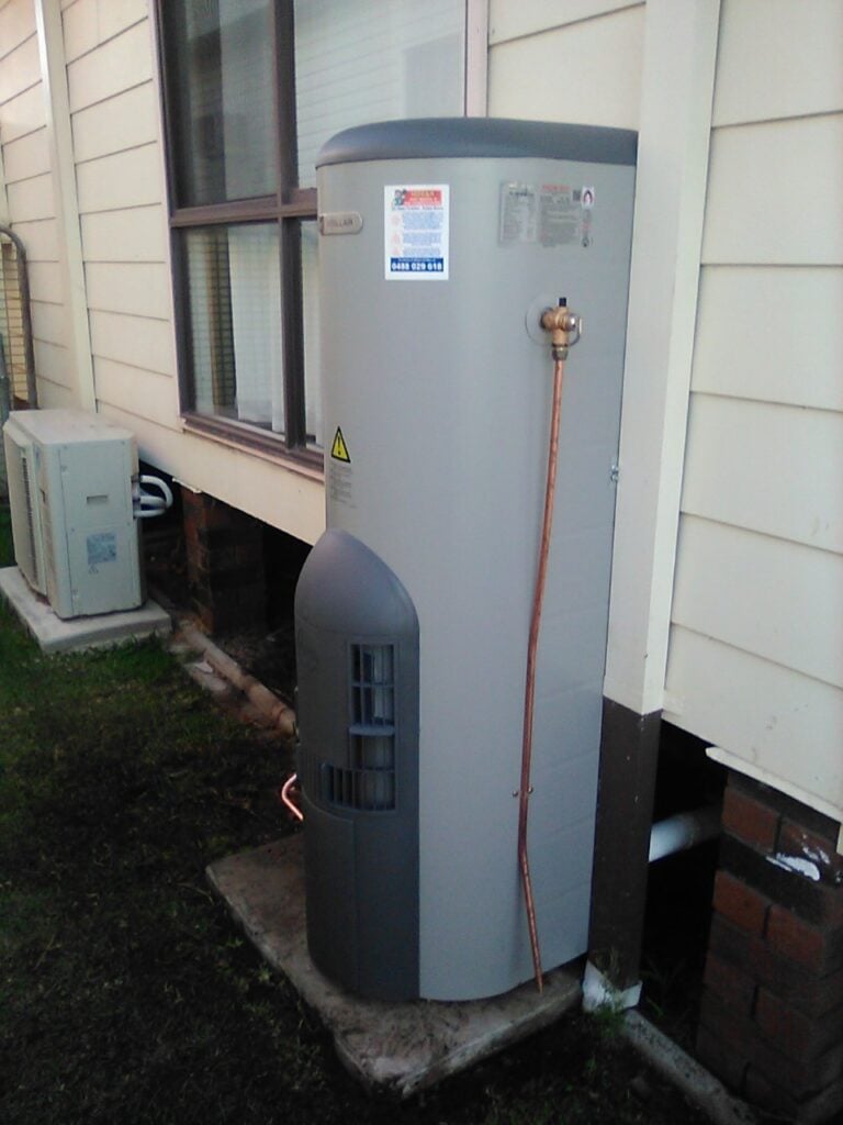 A tall, grey cylindrical heat pump water heater installed outdoors on a concrete slab next to a house with beige siding.