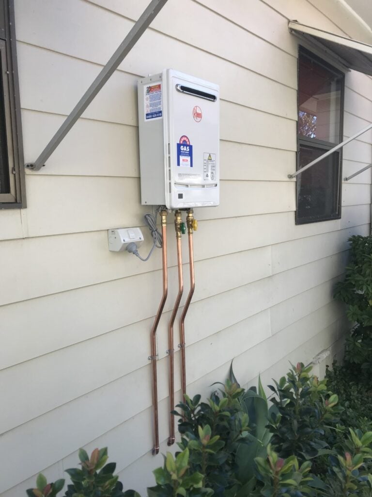 Wall-mounted white Rheem continuous flow gas hot water system with copper piping on a house exterior.