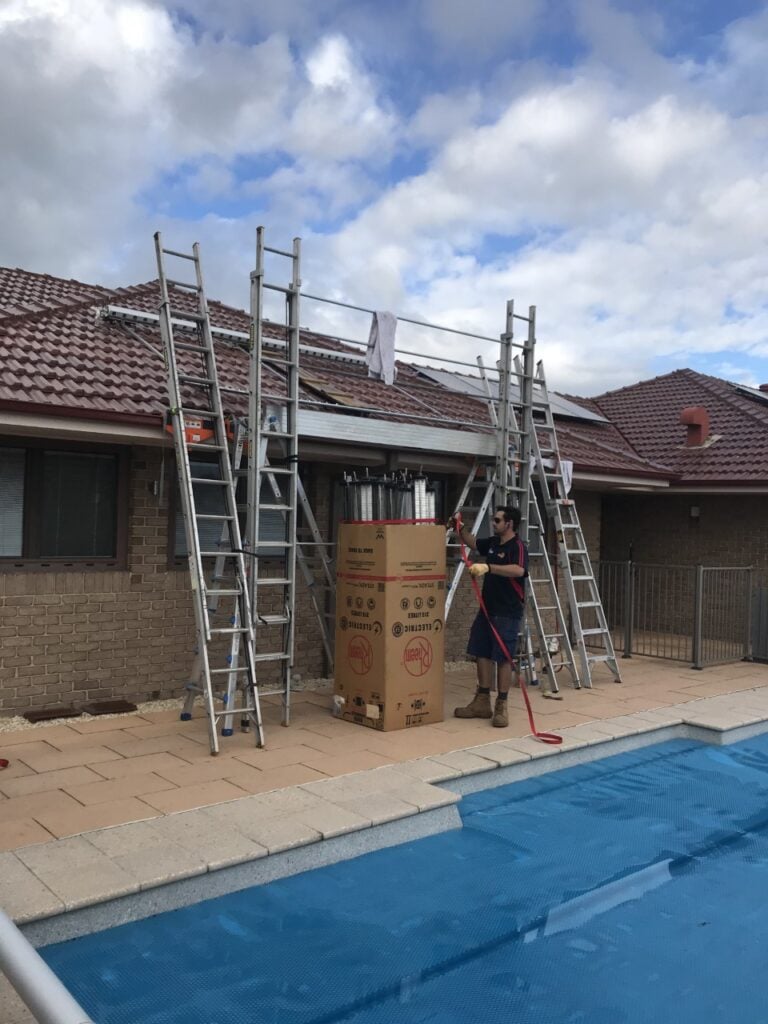 A technician unboxing a Rheem water heater next to a swimming pool, surrounded by extension ladders leaning against a roof.