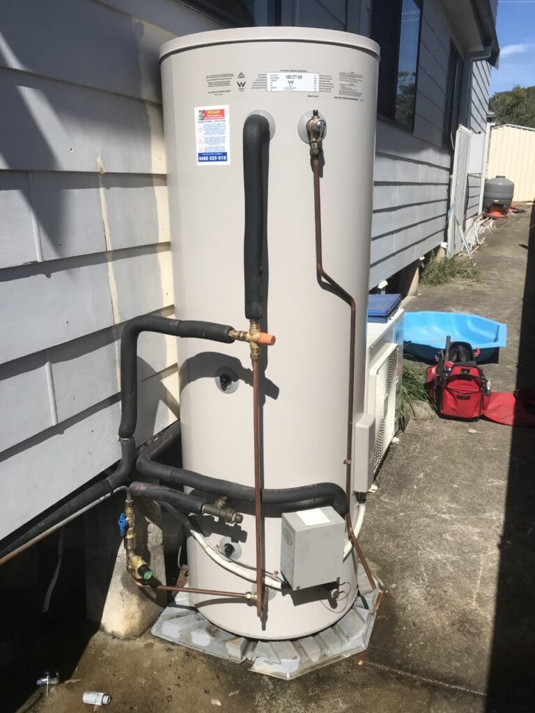 An outdoor electric storage hot water tank installed on a base alongside a weatherboard house wall with copper piping.