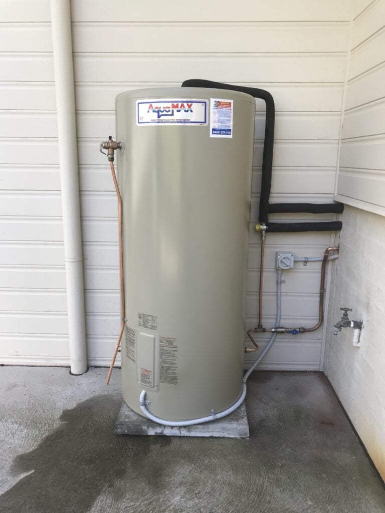 An AquaMax electric hot water heater installed on a concrete pad against an exterior wall.