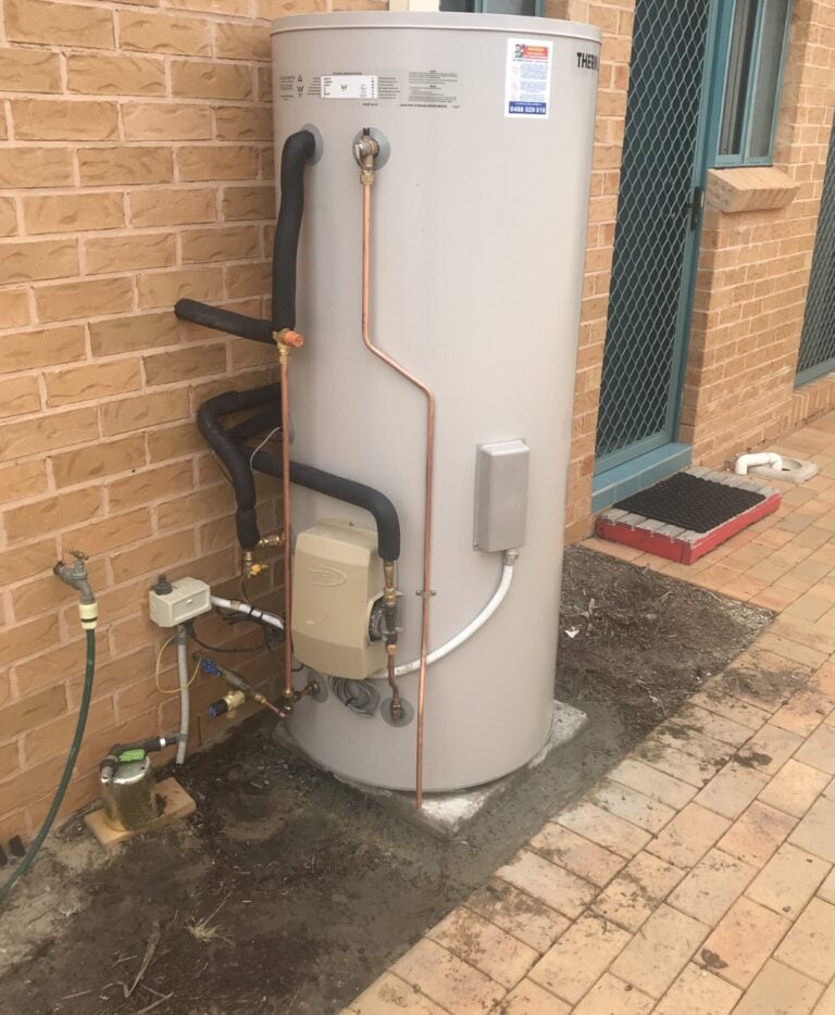 A Thermann hot water tank installed on a concrete slab against a brick house wall next to a paved walkway.
