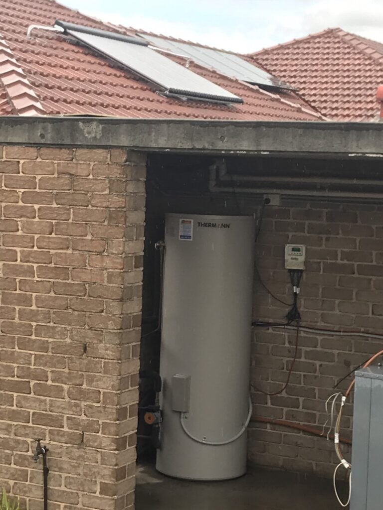 A Thermann solar hot water storage tank installed under an overhang, with solar panels visible on the tiled roof above.