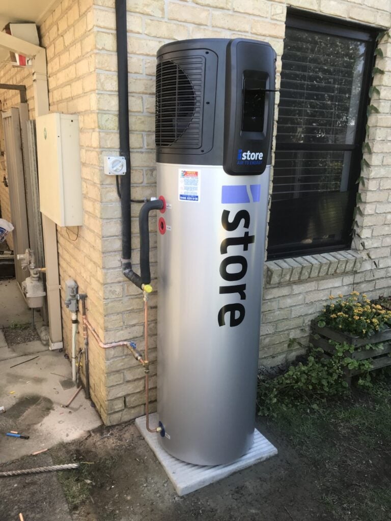 An outdoor installation of a silver iStore air-to-energy heat pump hot water system, positioned against a light brick wall next to a window.
