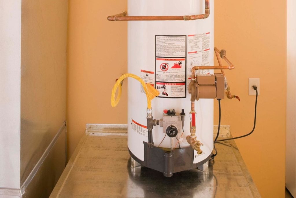 A white gas water heater tank with warning labels, a yellow flexible line, copper piping, and an attached circulation pump.