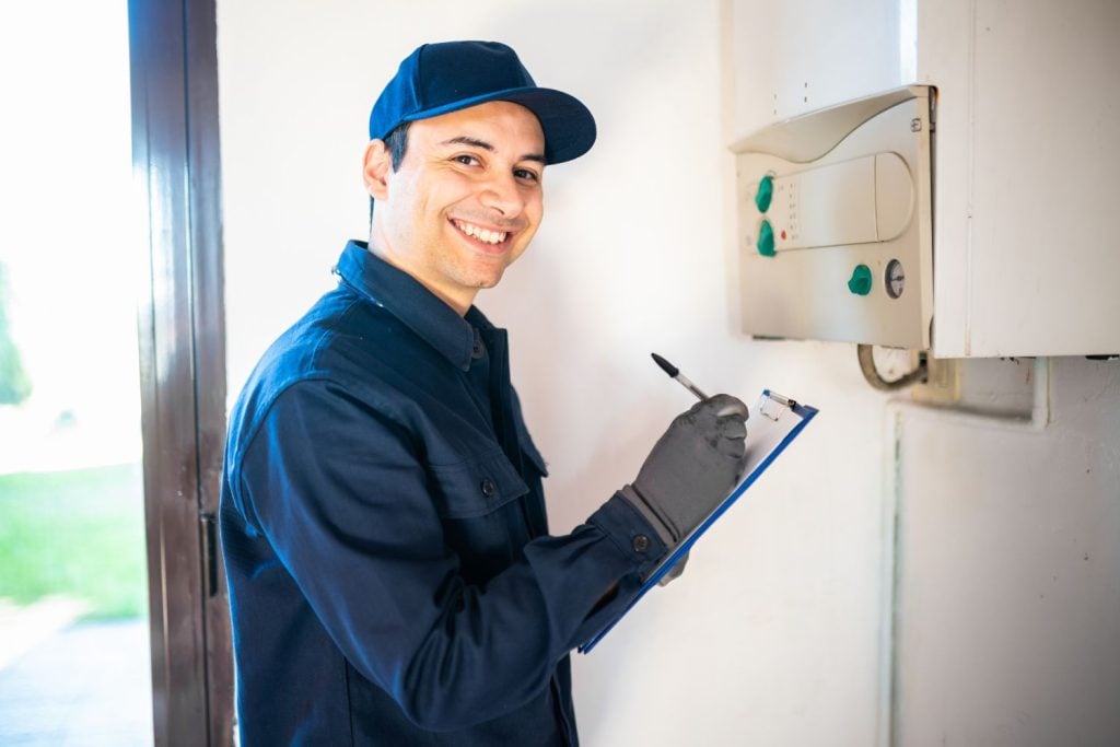 A smiling technician in a blue uniform and cap taking notes on a clipboard while standing next to a wall-mounted boiler.