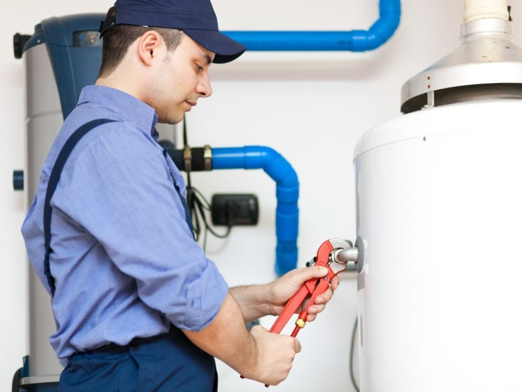 A technician wearing a dark blue cap, a light blue button-up shirt, and dark blue overalls using a red pipe wrench to adjust a metal valve pipe.
